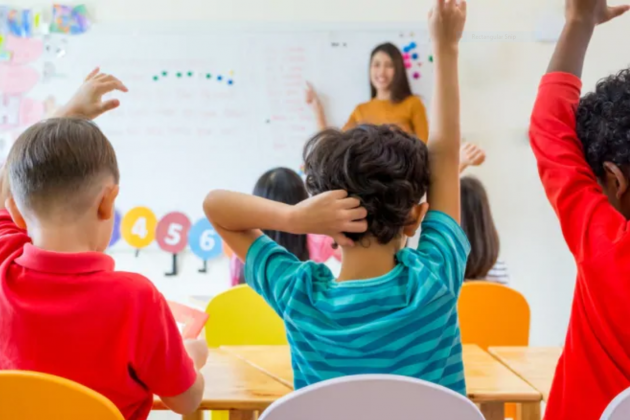 3 young boys raising their hands in a classroom facing a teacher at the front of the room